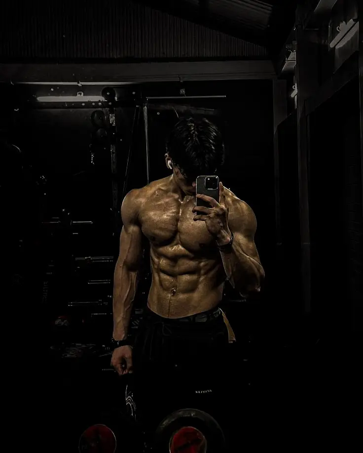 Shredded athletic man taking mirror selfie in dark gym showing defined abs and chest muscle definition